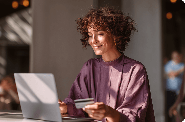 A smiling woman with curly brown hair, wearing a purple shirt, making an online payment with a credit card and laptop. Sunlight streams across the scene